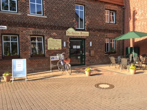Ein rotes Backsteingebäude, davor ein Werbeaufsteller, ein Fahrrad und eine Sitzmöglichkeit aus Gartenstühlen, Tischen und einem Sonnenschirm