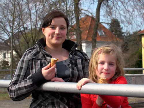 Eine Mutter und ihre Tochter, beider mit einer Eiswaffel in der Hand, stehen draußen an einem Geländer. Im Hintergrund sind einige Bäume ohne Grün zu sehen und ein weißes Wohnhaus mit spitzem roten Dach.