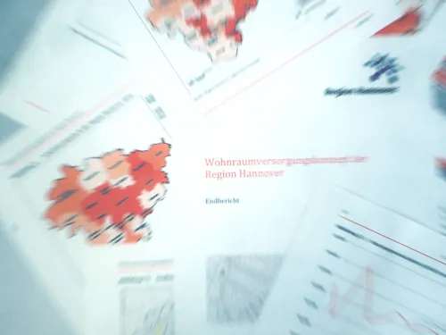 Deckblatt und einzelne Seiten des Wohnraumversorgungskonzeptes der Region Hannover liegen auf einem Tisch aus. Von einem zentralen Punkt aus wird die Szene dynamisch auseinandergerissen, sodass nur die Begriffe "Wohn" und "Region" scharf zu erkennen sind, nach außen hin nimmt die Unschärfe zu.