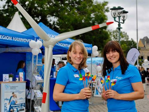 Zwei junge Frauen in blauen Poloshirts vor einem Miniaturwindrad und blauen Zelten