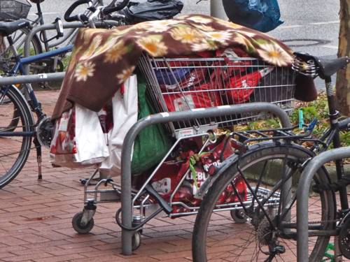 Ein Einkaufswagen mit Habseligkeiten parkt zwischen Zweirädern an einem Fahrradabstellbügel.