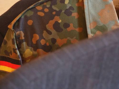 Auf einer Kleiderstange hängt ein Nadelstreifenanzug, dahinter ist eine Bundeswehr-Feldbluse in Tarnmuster.