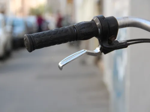 Zu sehen ist die Linke Seite eines Fahrradlenkers. Das Rad steht auf einem Gehweg und am Stra&szlig;enrand parken Autos.