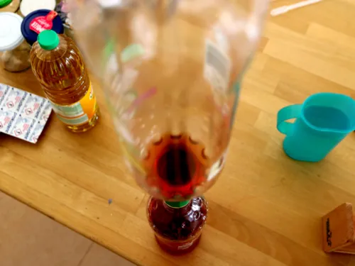 Eine rote Flüssigkeit fließt von einer PET-Flasche in eine darunter stehende. Auf dem Experimentiertisch sind weitere Utensilien, darunter eine Flasche mit Speiseöl.