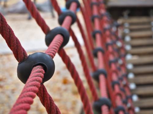 Rote Seile und Holzplanken bilden eine Hängebrücke und Geländer auf einem Spielplatz.