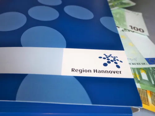 Aus einer blauen Mappe mit Regionslogo ragen Geldscheine hinaus.