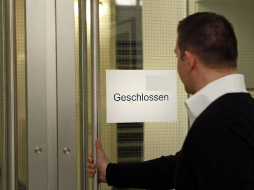 Ein Mann vor einer Glastür mit der rechten Hand am Türgriff. An der Tür ist ein Zettel mit dem Hinweis "geschlossen" befestigt.
