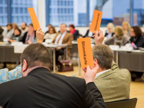 Mitglieder der Regionsversammlung heben orangefarbene Pappkarten mit dem schwarzen Text "Stimmkarte" in die Luft.
