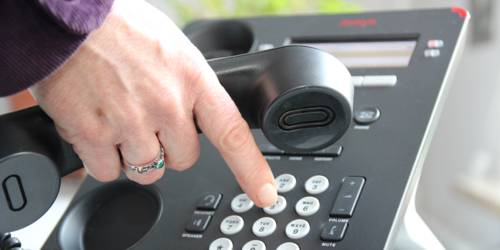 Graues Telefon, eine Hand, die den Hörer hält und gleichzeitig am Wählen einer Nummer ist