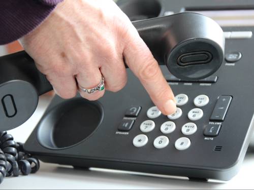 Graues Telefon, eine Hand, die den Hörer hält und gleichzeitig am Wählen einer Nummer ist