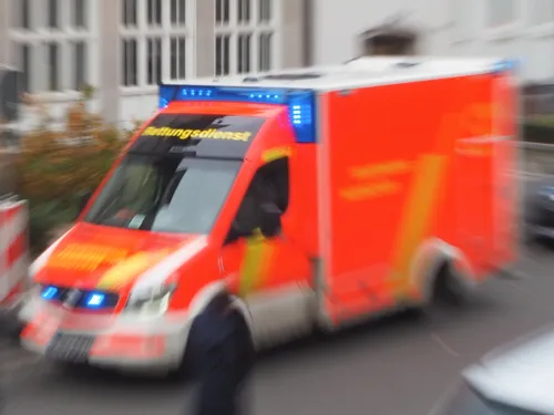 Ein Rettungswagen steht auf einer Straße, blaue Rundumkennleuchten leuchten auf. Durch den Fotoeffekt Reisszoom wirkt das Bild dynamisch. Menschen queren die Straße vor dem Rettungswagen.