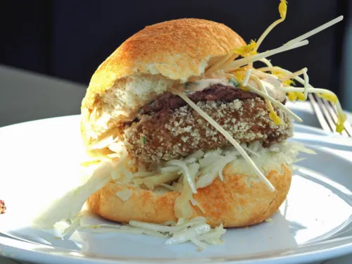 Zwischen zwei hellen, knusprigen Brötchenhälften sind Krautsalat, eine vegetarische Bulette, Rotkrautdip, Knoblauchsoße und Sprossen übereinander gestapelt. Der vegetarische Burger liegt auf einem Teller und wird von links von Sonnenlicht beschienen. Im Hintergrund liegt eine Gabel.