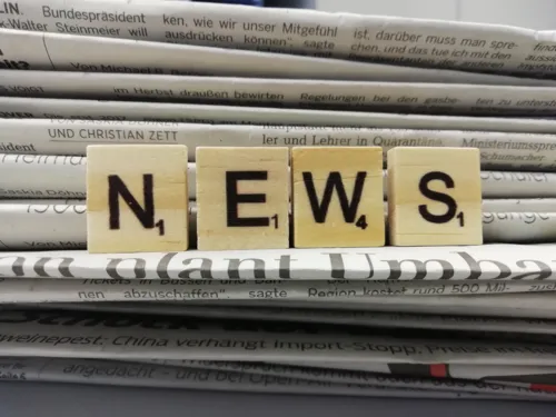 Vier Holzwürfel mit Buchstaben drauf, die nebeneinander aufgestellt das Wort "News" ergeben. Dahinter ist ein Stapel Zeitungen zu sehen.