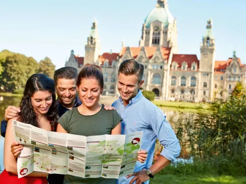 Vier junge Erwachsene betrachten eine Stadtkarte vor demhistorischen Neuen Rathaus mit grüner Kuppel.