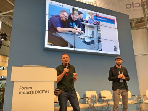 Zwei Männer mit Mikrofonen stehen auf einer Bühne in einer Messehalle. Hinter ihnen läuft auf einem großen Bildschirm eine PowerPoint Präsentation zum Thema Digitale Inklusion - wie Menschen mit Einschränkungen neue Medien nutzen.