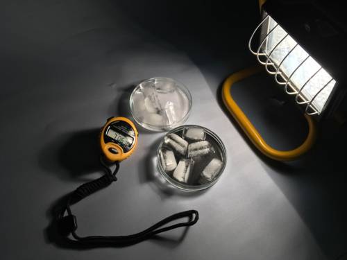 Eine Versuchsanordnung, bei der eine stark Wärme erzeugende Lampe an zwei mit Eiswürfel gefüllte Schalen gerückt wurde, daneben eine Stoppuhr, um die Zeit zu messen, bis die Eiswürfel durch die Wärme zu Wasser geworden sind.
