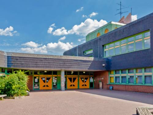 Der Eingangsbereich des Schulzentrums Bothfeld: Drei orange doppelseitige Schwingtüren und grüne Fensterelemente in einem Flachbau, der unten rot verklinkert und oben mit schwarzen Schindeln versehen ist.