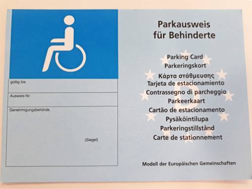 Der Parkausweis für Behinderte zeigt links das Symbol für eine Person mit Rollstuhl, darunter die Felder "Gültigkeit, Ausweisnummer und Genehmigungsbehörde". Rechts befinden sich unterhalb der Worte "Parkausweis für Behinderte" Übersetzungen des Begriffs in anderen Sprachen. 