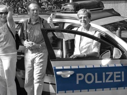 Drei Personen (zwei Frauen und ein Mann) stehen lächelnd vor einem Polizeifahrzeug. Die beiden Frauen halten den Daumen nach oben. Das Foto ist schwarz-weiß, nur die Beschriftung der Polizei ist blau eingefärbt.