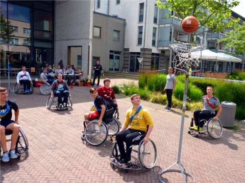 Auf dem Bild sind acht junge Rollstuhlfahrer zu sehen, die auf einem extra hergerichteten Platz mit zwei mobilen Basketballkörben Basketball spielen.