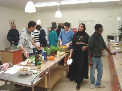 Mehrere Personen stehen in einer Wohnküche und kochen gemeinsam. Jeder macht etwas anderers. Ein Mann rührt etwas zusammen, ein Mann reicht etwas rüber. Einige unterhalten sich und andere gehen derzeit durch die Wohnküche.