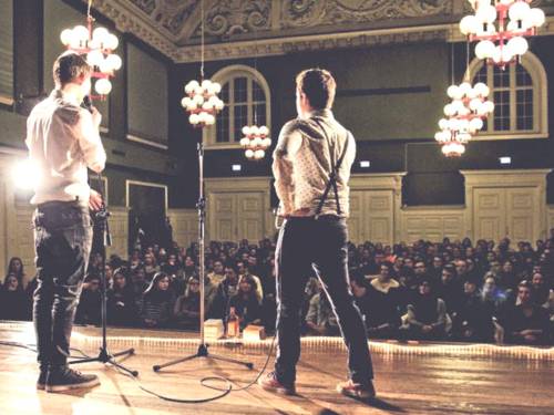 Zu sehen sind zwei Akteure auf der Bühne mit Blick in den voll besetzten Saal des Lister Turm