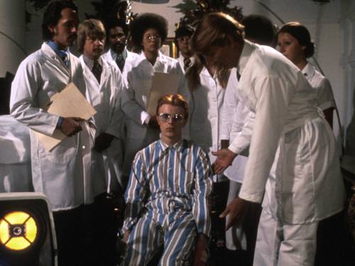 Im Bild zu sehen ist David Bowie, der zwischen einer Gruppe Ärzte sitzt und mit leeren Blick ins Nichts starrt.