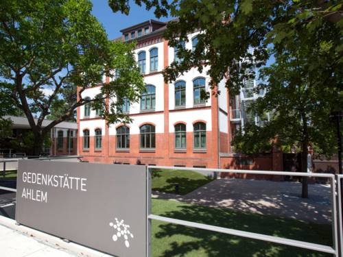 Von der Heisterbergalle aus gesehen: Im Vordergrund ist ein Metallschild mit Aufschrift "Gedenkstätte Ahlem" und Regionslogo, dahinter steht das Gebäude der Gedenkstätte Ahlem. 