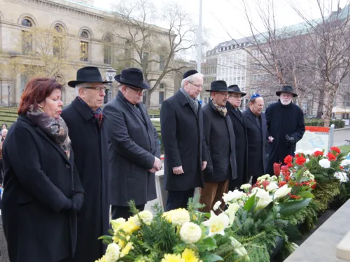 Menschen in Gedenken vor einem Mahnmal voller Blumen. 