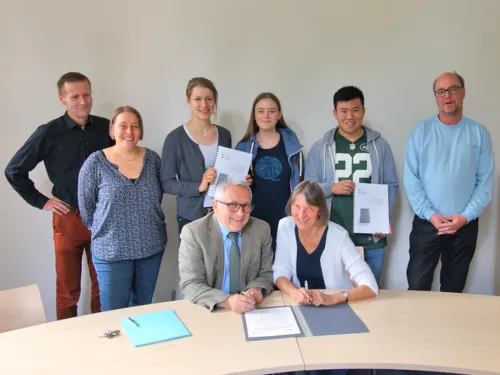 Zwei Personen sitzen an einem Tisch und unterzeichnen ein Dokument, dahinter stehen sechs weitere Personen