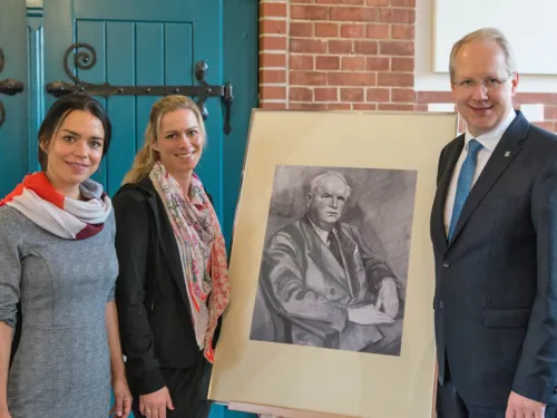 Drei Personen neben einer Staffelei, auf der ein Portrait eines Mannes steht