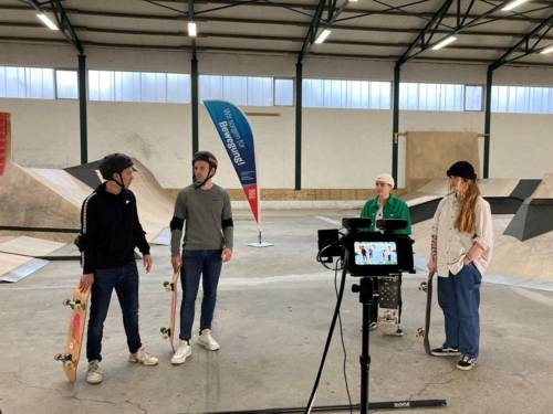 Vier Personen mit Skateboards in einer Skatehalle. Sie stehen vor einer Kamera. 