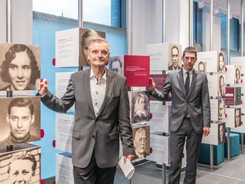 Zwei Männer stehen an zwei Säulen, auf denen Porträtfotos von anderen Menschen zu sehen sind