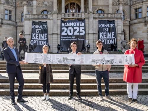 Fünf Personen stehen vor dem Neuen Rathaus in Hannover und präsentieren einen Teil des zweiten Bid Books zur Bewerbung als Kulturhauptstadt Europas 2025