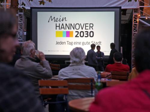 Symbolbild für den liveStream- Menschen schauen auf einer Leinwand den Trailer "Mein Hannover 2030"