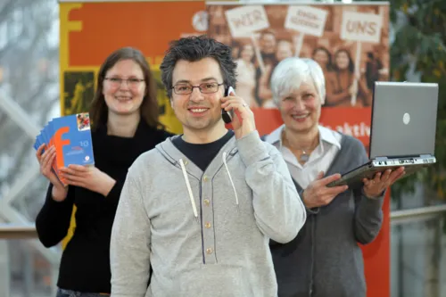 Ein junger Mann lächelt mit einem Telefon am Ohr ins Bild. Hinter ihm stehen rechts eine ältere Frau mit einem Laptop und links eine jüngere Frau mit Flyern in der Hand.
