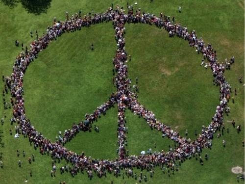Menschen haben sich auf einer Grünfläche zum internationalen Friedenssymbol (Kreis mit mittigem Trennstrich und zwei Seitenarmen) aufgestellt.