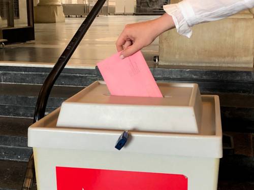Ein Briefumschlag wird in eine Wahlurne gesteckt (nachgestellte Szene).