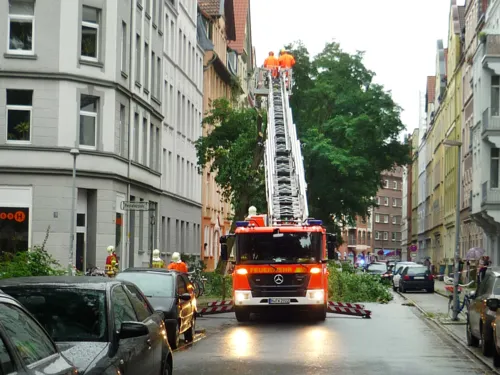 Feuerwehrfahrzeug in einer engen Straße. Der umzustürzen drohende Baum wird von Einsatzkräften der Feuerwehr begutachtet.