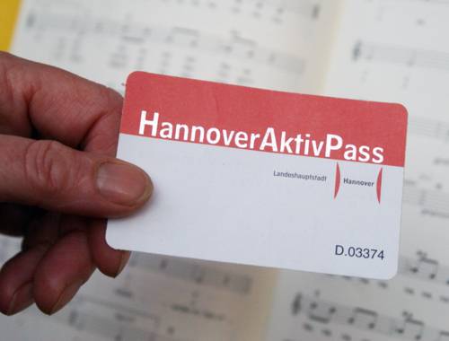 Im Vordergrund ist eine Hand zu sehen, die den rot-weißen Hannover-Aktiv-Pass hält, im Hintergrund liegen Notenblätter