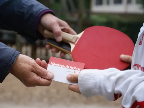 Die H&auml;nde von einem Erwachsenem und einem Kind tauschen den Hannover-Aktiv-Pass und zwei Tischtennisschl&auml;ger aus