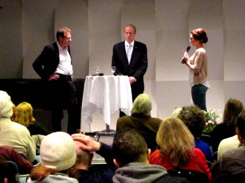 Drei Personen stehen auf einer Bühne an einem runden Stehtisch, die Moderatorin rechts hält ein Mikrofon in der Hand und spricht den beiden Männern zugewandt. Unten im Bild sieht man einen Teil der Zuhörer/innen von hinten.