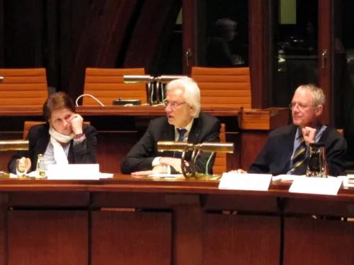 Im Hodlersaal sitzen drei Personen in einer Reihe. Die Linke stützt den Kopf ab und bickt nach rechts, die Mittlere redet, die Rechte schaut leicht nach links. Vor den Personen stehen Namensschilder und eine Tischlampe mit zwei fest verbauten Mikrofonen.