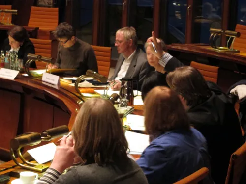 Acht Personen sitzen im Viertelkreis vor Pulten. Einer (rechts im Bild) gibt ein Handzeichen.