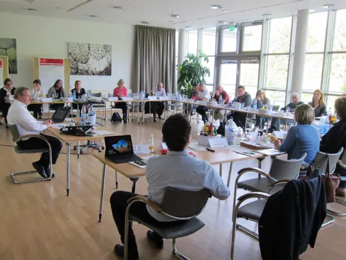 Etwa 25 Personen sitzen in einem Diskussionssaal hinter viereckig aufgestellten Tischen. Einige davon gestikulieren. Auf den Tischen liegen Papiere, Laptops und Getränke.