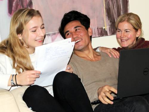 Zwei Jugendliche und eine Frau sitzen nebeneinander auf einem Sofa. Die Jugendliche links hält einen Stapel Papiere in der Hand, der Jugendliche in der Mitte hat einen laptop auf dem Schoß. Die Frau rechts im Bild blickt zum Jugendlichen in der Mitte. Alle drei lächeln.