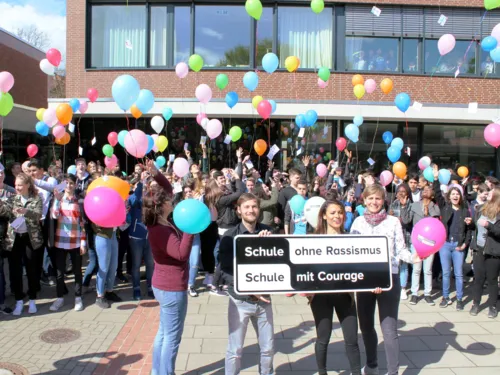 Etwa 80 Sch&uuml;ler/innen stehen hinter vier Lehrer/innen die das typische SOR-SMC-Banner vor sich halten. Die Sch&uuml;ler/innen lassen Ballons in den Himmel steigen, an denen Postkarten befestigt sind.