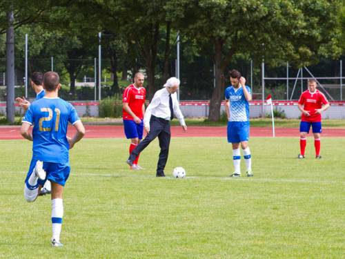 Ein Mann in Hemd und Anzughose führt einen Anstoß beim Fußball aus, um ihn herum Spieler in roten und blauen Trikots