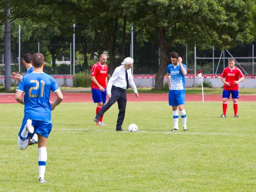 Ein Mann in Hemd und Anzughose führt einen Anstoß beim Fußball aus, um ihn herum Spieler in roten und blauen Trikots