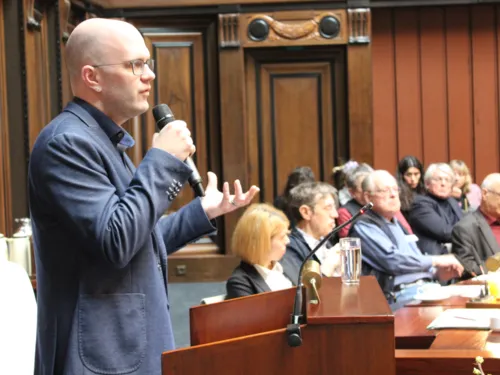 Ein Mann steht im Hodlersaal des Neuen Rathauses hinter einem Stehpult mit einem Mikrofon in der Hand und spricht. Rechts im Bild sind sitzende Zuhörer*innen erkennbar.